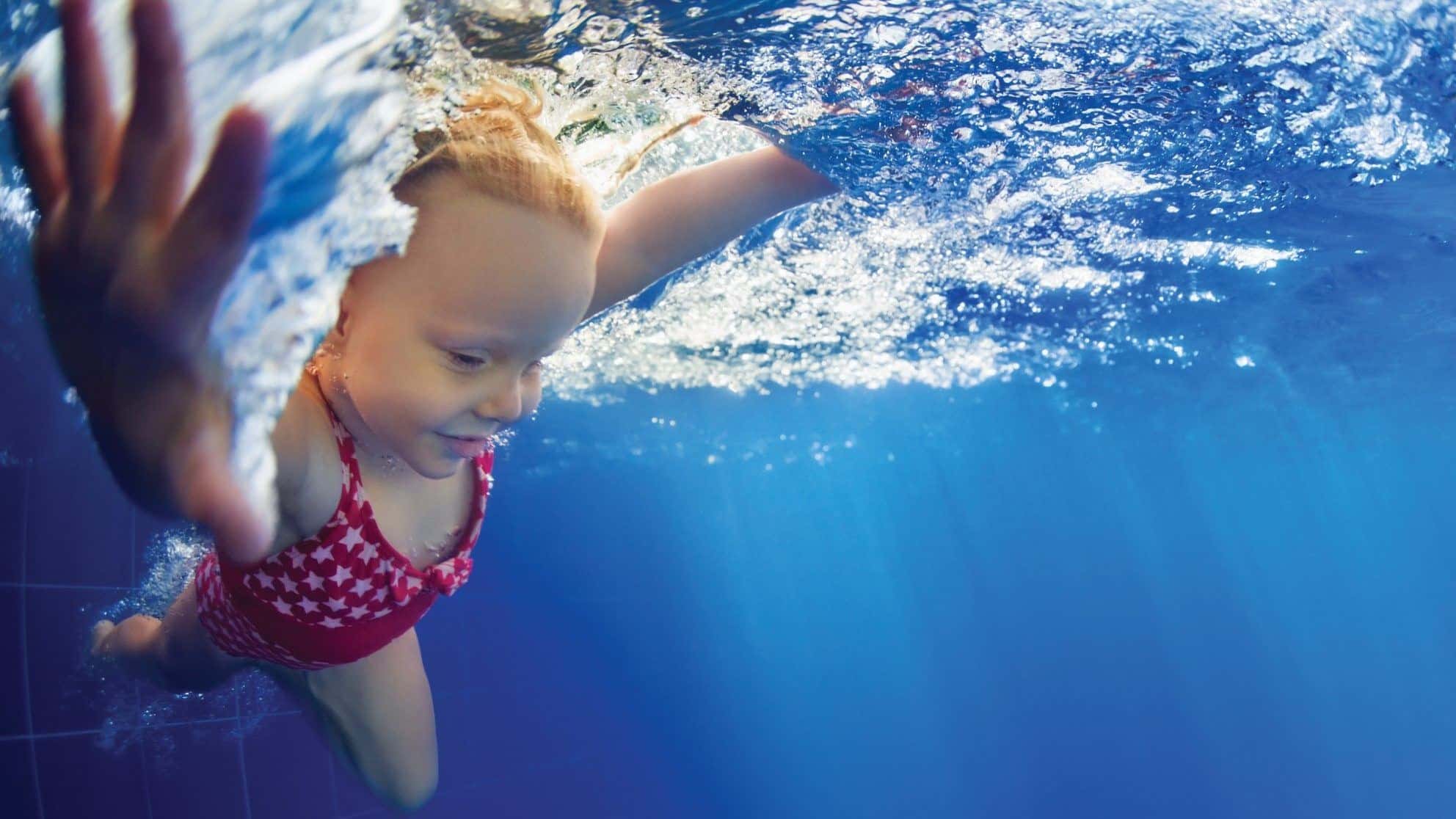 A young girl swims underwater with her eyes open in a clear blue pool