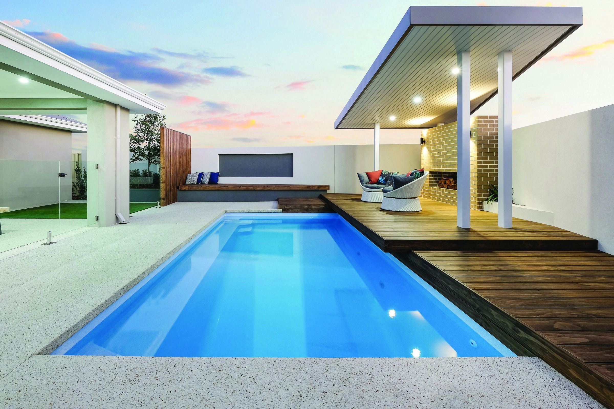 A long blue lap pool beside a modern home with a covered outdoor lounge at dusk