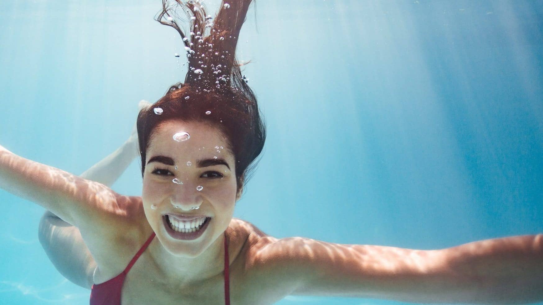 A smiling woman swims underwater with her eyes open in a freshwater pool