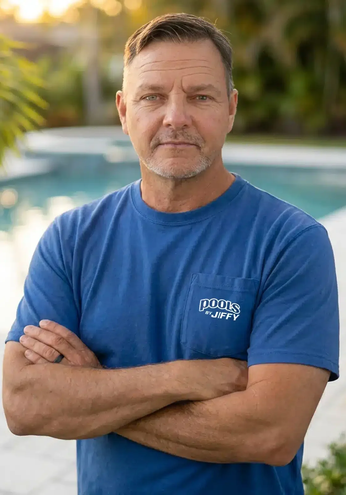 A man in a blue Pools by Jiffy T-shirt stands with arms crossed by a swimming pool, looking at the camera. Trees and sunlight are visible in the background.