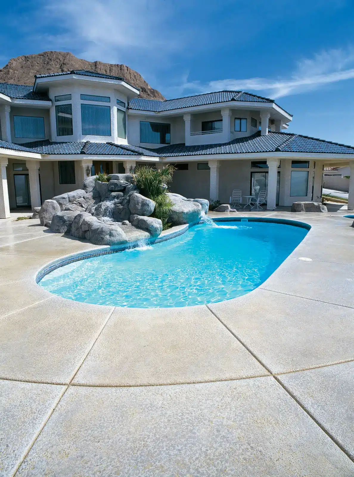 Modern house with a curved swimming pool, rock waterfall feature, and patio area under a clear blue sky with mountains in the background.