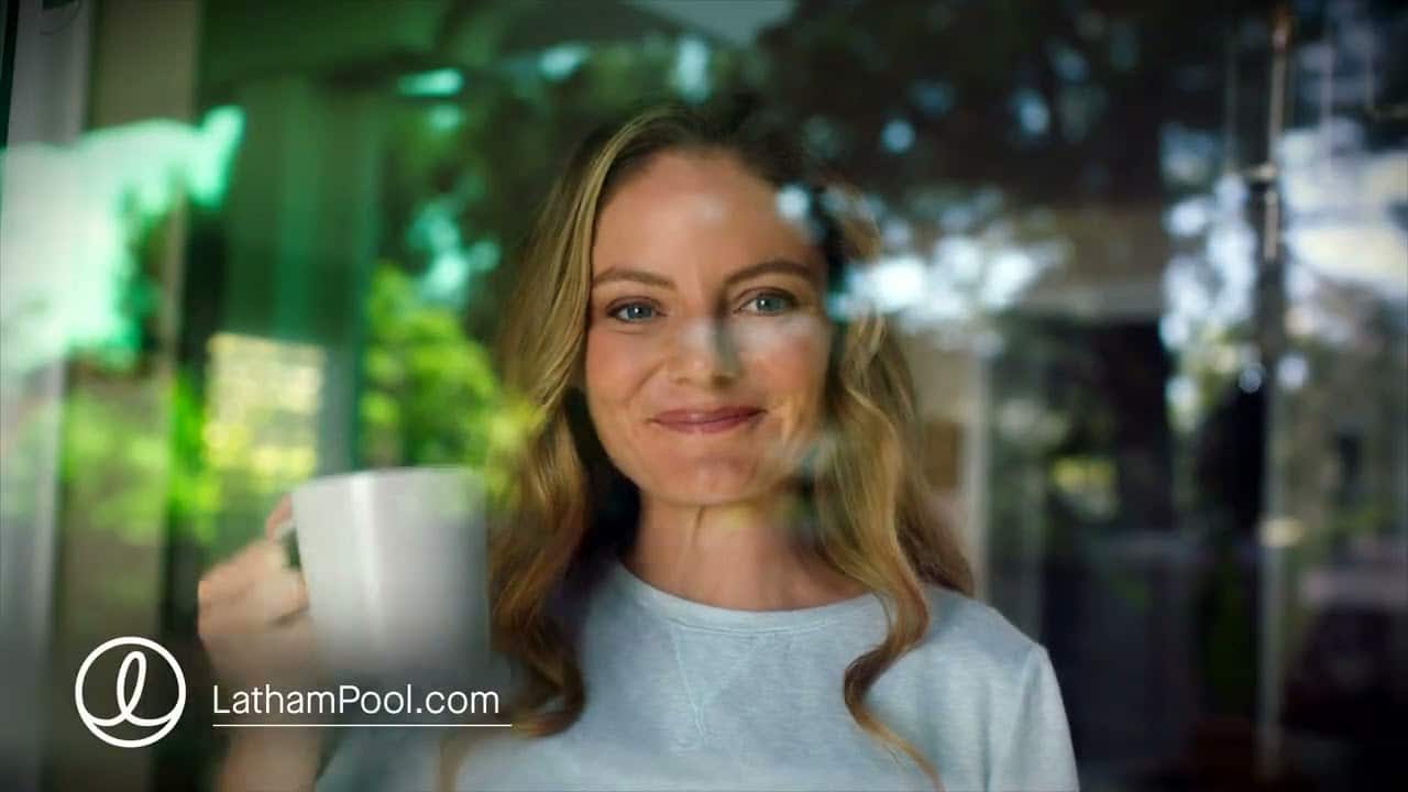 Woman with wavy hair smiles and holds a white mug while standing by a window. Greenery and reflections are visible outside. The logo and text LathamPool.com appear in the lower left corner.