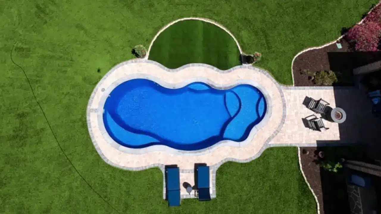 Aerial view of a uniquely shaped blue swimming pool with a stone deck, two lounge chairs at one end, surrounded by green grass, and a patio dining set nearby.