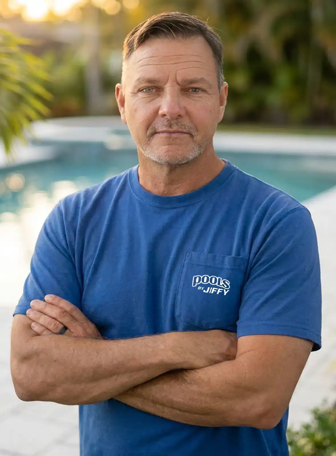 A man in a blue Pools by Jiffy T-shirt stands with arms crossed by a swimming pool, looking at the camera. Trees and sunlight are visible in the background.