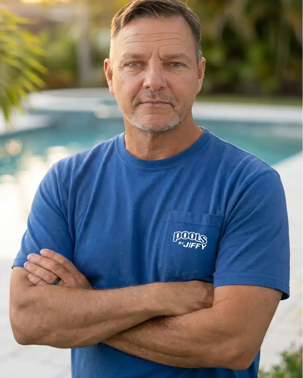 A middle-aged man with short hair stands with arms crossed, wearing a blue Pools by Jiffy T-shirt, near a pool with greenery in the background—embodying the confidence and experience of a trusted pool contractor in Palm Coast.