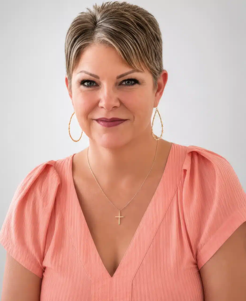 A woman with short, light brown hair wearing a peach blouse, gold hoop earrings, and a cross necklace smiles at the camera against a plain light background—her confident demeanor reflects the trusted professionalism you’d expect from a leading pool contractor Palm Coast residents rely on.