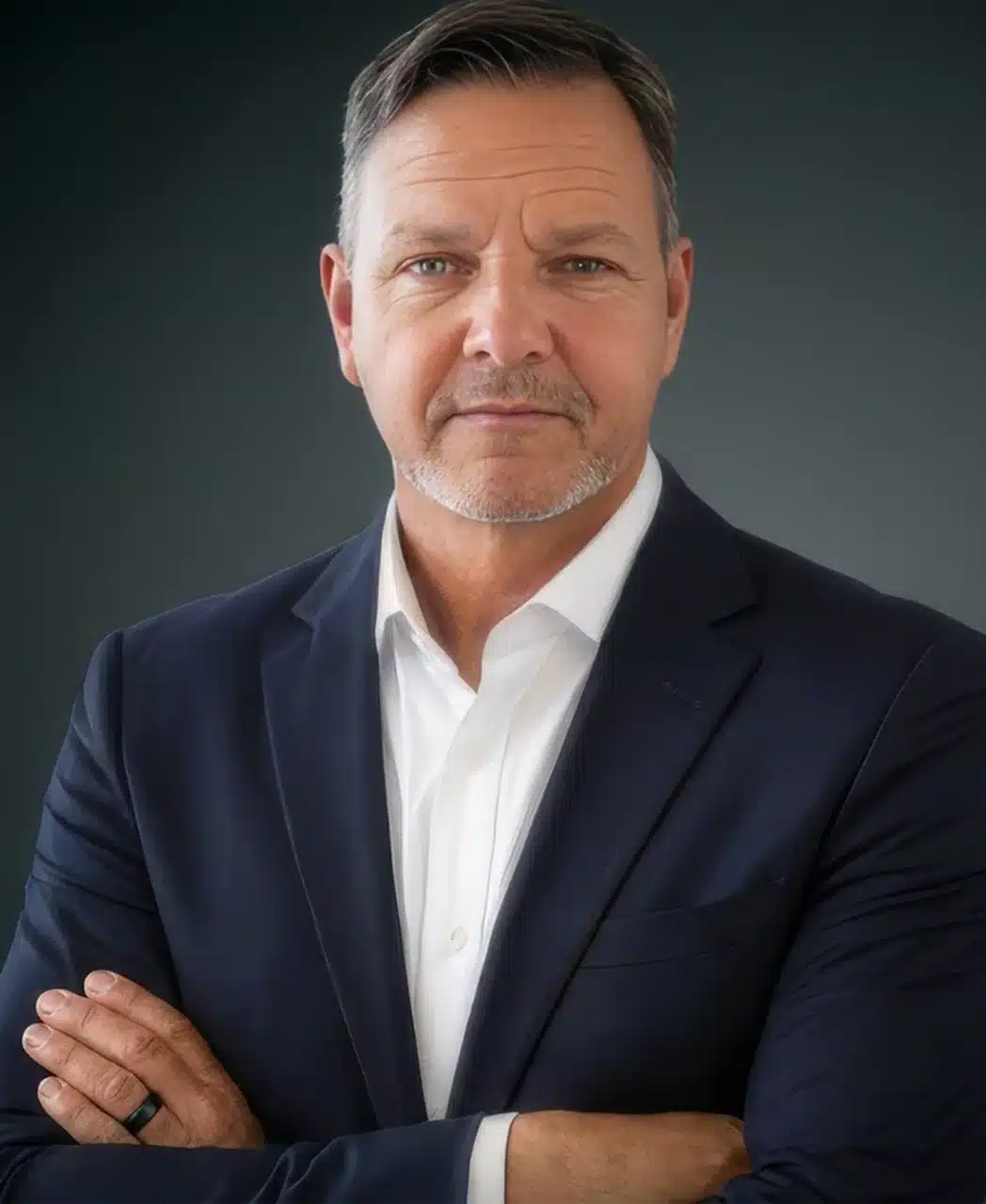 Middle-aged man with short brown hair and a trimmed gray beard, wearing a dark suit jacket and white dress shirt, stands against a dark background with arms crossed and a serious expression, embodying the professionalism expected of a leading pool contractor in Palm Coast.